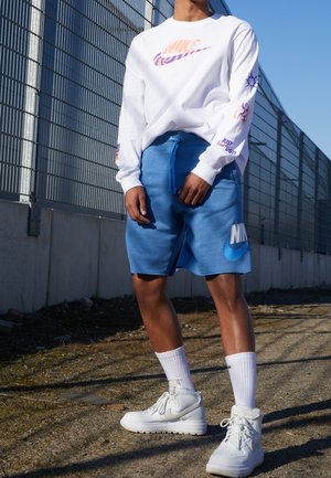 Hvid Nike langærmet trøje med farverigt logo, blå shorts med stort Nike-logo, hvide høje sneakers og hvide mellemlange sokker.
