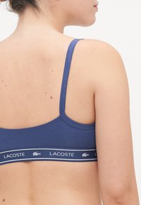 Blå bomulls-bh med justerbare stropper og en merkevare-bånd med hvit logo og detaljer. Teksturen ser glatt og tettsittende ut.