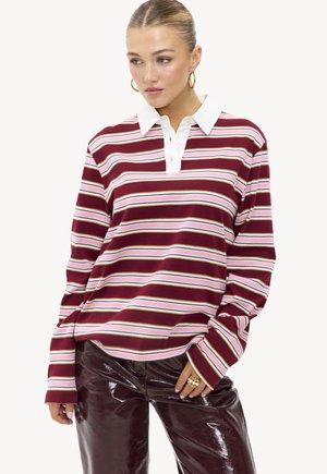Femme portant un polo rayé bordeaux, rose et blanc à manches longues avec un col blanc et un pantalon bordeaux brillant, les mains détendues.