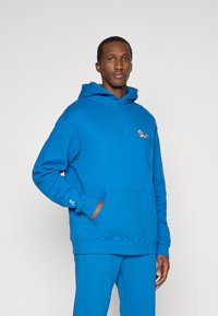 Μπλε πουλόβερ φούτερ από μαλακό ύφασμα, με καγκουρό τσέπη, ραμμένες μανσέτες και μια μικρή γραφική λεπτομέρεια στο στήθος.