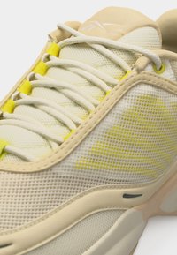 Baskets en mesh beige et jaune avec lacets blancs, languette rembourrée et semelle épaisse, présentées en gros plan.