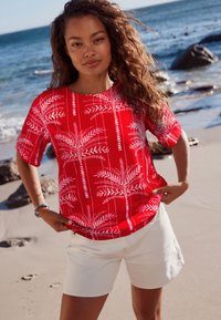 Rotes Kurzarmhemd mit weißem Palmenblattmuster, kombiniert mit weißen Shorts. Verfügt über eine lockere Passform und einen runden Ausschnitt.