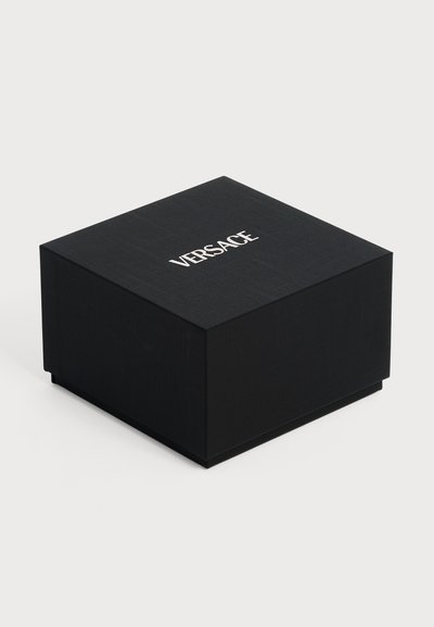 Boîte carrée noire avec surface texturée et logo Versace blanc centré en haut, présentée sur un fond clair uni.