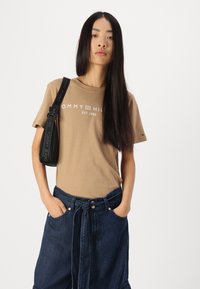 Brūna īsām piedurknēm T-kreklu ar balto "TOMMY HILFIGER EST. 1985" logotipu. Apvienots ar tumši zilu džinsu svārku, kuram ir aukla jostas vietā un priekšējās kabatas.