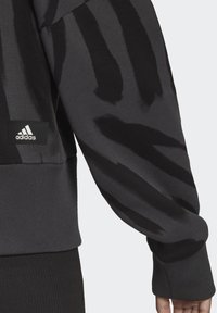 Felpa grigia scura con strisce astraubenere, polsini a costine e logo Adidas su un'etichetta nera vicino all'orlo. Texture morbida.