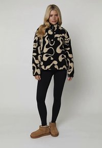 Pull-over en polaire noir et beige avec un motif de tourbillons abstraits, col zippé et manches longues, associé à des leggings noirs et des bottes beiges.