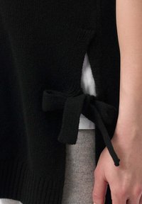 Svart stickad tröja med sidoflikar, med en strukturerad tygkvalitet och en knytning i midjan. Lagd över en ljusgrå topp.