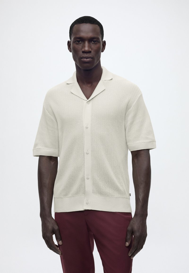 Άνδρας που φοράει μπλούζα beige με κοντά μανίκια, κουμπιά, πλεκτή ύφανση, γιακά και μπορντό παντελόνι, στέκεται μπροστά σε ένα απλό λευκό φόντο.