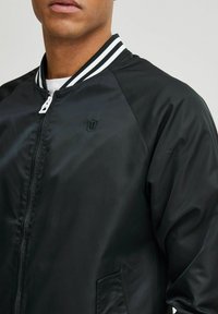 Giubbotto bomber nero con una texture liscia e lucida. Presenta un colletto a righe bianche, chiusura con zip e un piccolo logo sul lato sinistro del petto.