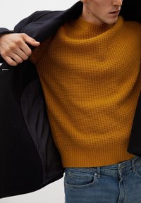 Black coat with a navy lining, worn over a textured mustard yellow sweater. Sweater features a ribbed hem, paired with blue jeans.