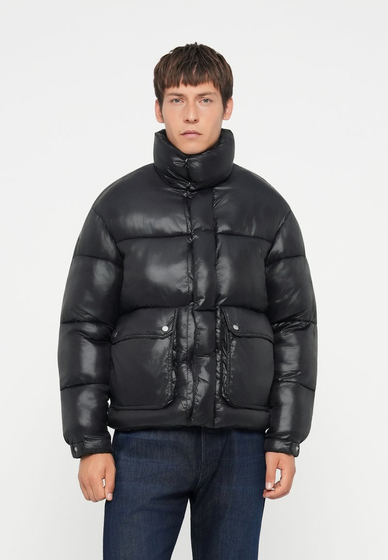 Schwarze Steppjacke mit hohem Kragen, glänzender Oberfläche, horizontaler Steppung, zwei Fronttaschen und Druckknopfverschlüssen. Über Denim getragen.