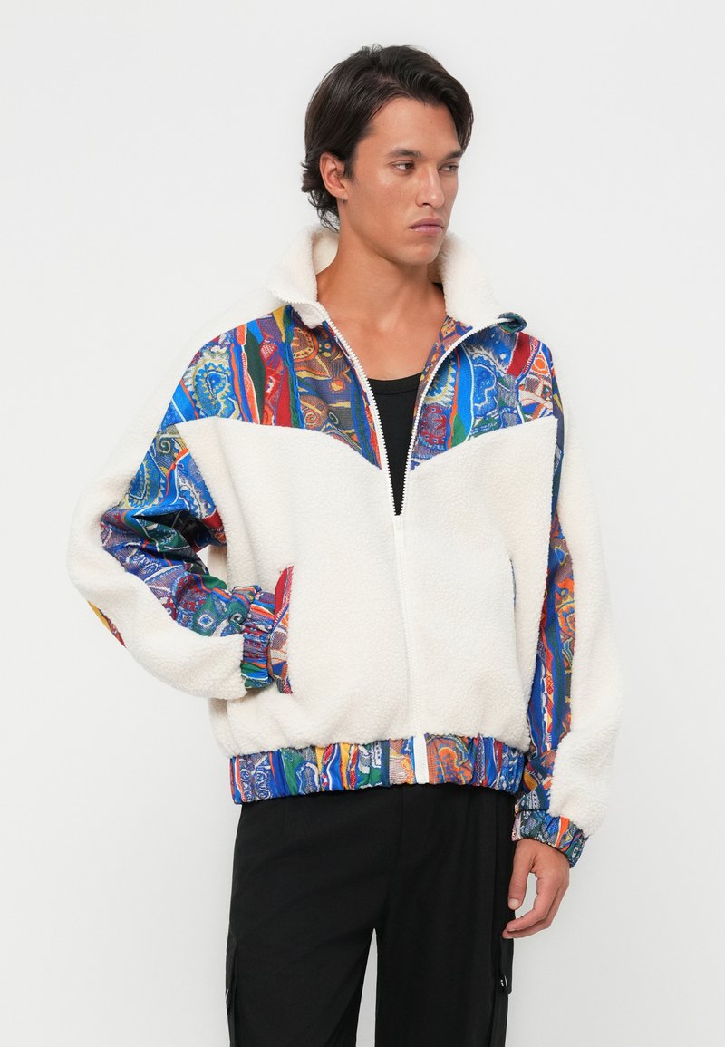 Chaqueta de forro polar blanca con tela de patrón colorido en los hombros y las mangas. Cierre de cremallera en el frente, cuello alto, puños elásticos y dobladillo. Camiseta negra debajo.