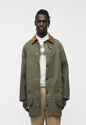 Młody mężczyzna w oliwkowo-zielonej kurtce z brązowym kołnierzem, białej polo w wzory i beżowych spodniach, stojący na tle jednolitego tła.