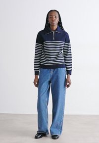 Pullover a righe blu navy e bianche in maglia con colletto zip, abbinato a jeans blu a vita alta e scarpe nere. Sfondo semplice.