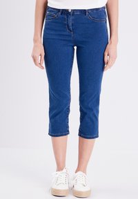 Blauwe denim capri jeans met een getailleerd ontwerp, een gemiddelde taillehoogte, vier zakken en een zoom die halverwege de kuit komt. Gecombineerd met witte sneakers.