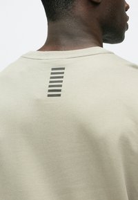 Ανοιχτό μπεζ μπλουζάκι με στρογγυλή λαιμόκοψη, που φέρει κάθετη μαύρη γραμμή στο πίσω μέρος. Απαλό βαμβακερό υλικό, casual εφαρμογή.