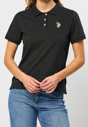 Kobieta ubrana w czarną koszulkę polo z krótkim rękawem, trzema guzikami i logo na piersi, połączoną z niebieskimi dżinsami, ręce złączone z przodu.