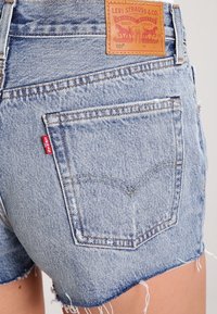 Denimshorts i en urblekt blå färg, fransad fåll och två bakfickor, märkesskylt på midjebandet, med den ikoniska röda Levi's-fliken.