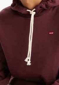 Bordová mikina s kapucí vyrobená z měkké tkaniny, která má šňůrku na stahování s krémovými provázky a malým červeným logem Levi's na přední straně.