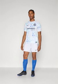 Jaunas vyras sportininkas, vilkintis baltais ir mėlynais raštais futbolo marškiniais, balta apranga, mėlynomis kojinėmis ir juodais bateliais, stovi prieš vienodą foną.