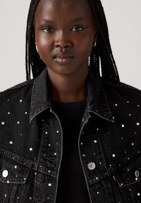 Zwarte denimjas met verspreide zilveren studs, klassieke kraag, knoopsluitingen en borstzakken. Gelaagde stof voegt detail toe.