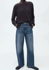 Pull en maille foncée à coupe ample avec épaules tombantes, associé à un jean bleu taille haute et longueur complète, et des chaussures noires slip-on.