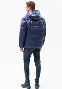 Marineblauwe pufferjack met een glanzende afwerking, kap en sectieontwerp. Draagt het in combinatie met donkere spijkerbroeken en laarzen, wat een casual stijl laat zien.