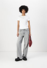 Maglietta bianca a coste con maniche corte e un piccolo logo, abbinata a pantaloni jeans grigi larghi e scarpe con plateau nere. Tiene una borsa rossa a tracolla.