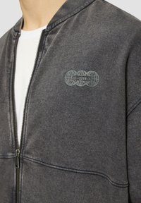 Graue Zip-Jacke mit strukturiertem Stoff, geripptem Kragen und dezentem Logo auf der linken Brust. Bietet ein schlankes Design und einen funktionalen Reißverschluss.