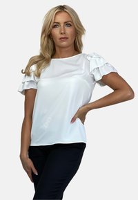 Biała bluzka z krótkimi, falbankowymi rękawami, wykonana z gładkiego materiału. Charakteryzuje się luźnym krojem i okrągłym dekoltem, zestawiona z czarnymi spodniami.