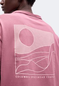 Άτομο που φοράει ροζ μπλούζα Columbia Sportswear με λευκό αφηρημένο σχεδιασμό γραμμών από λόφους και ήλιο στην πλάτη.