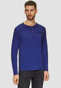 Pitkähihaisessa sinisessä henley-paidassa on teksturoitu pinta, jossa on napitettava kaulus ja hillitty musta teksti rinnassa.