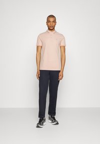 Kurzärmliges Poloshirt in Hellrosa mit kontrastierendem Kragenbesatz, kombiniert mit marineblauen Hosen und schwarzen Turnschuhen, vor einem einfarbigen Hintergrund stehend.
