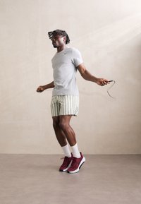 Gray athletic t-shirt, green and white striped shorts, white socks, and red Nike sneakers; person skipping rope, mid-action against a neutral backdrop.