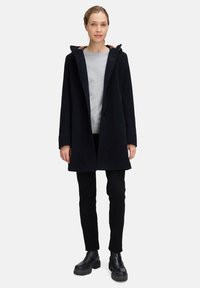 Vrouw met blond haar dat teruggebonden is, gekleed in een zwart gewatteerd jasje met capuchon, een grijze trui, zwarte broek en zwarte chunky laarzen, staand tegen een witte achtergrond.