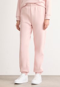 Ροζ, ελαφριά, tapered φόρμες με ελαστική μέση και μανσέτες στους αστραγάλους, συνδυασμένες με λευκά αθλητικά παπούτσια σε ουδέτερη φόντο.