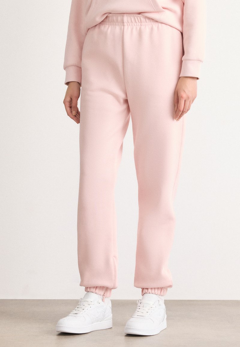 Rosa, lette, tapered joggebukser med elastisk midjebånd og mansjetterte ankler, kombinert med hvite sneakers på en nøytral bakgrunn.