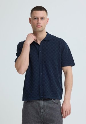 Giovane uomo che indossa una camicia blu navy a maniche corte con texture a scacchi e jeans grigi, in piedi contro uno sfondo chiaro e semplice.