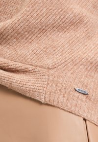 Hellrosa, gestrickter Pullover mit einem strukturierten Muster, ausgestattet mit einem kleinen silbernen Logoschild. Der Stoff wirkt weich und gemütlich vor einem beigen Hintergrund.