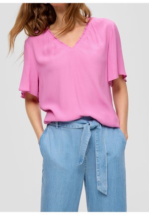 Frau trägt eine lockere, rosa V-Ausschnitt-Bluse und hellblaue, hoch taillierte Jeans mit gebundenem Gürtel, Hände in den Taschen.