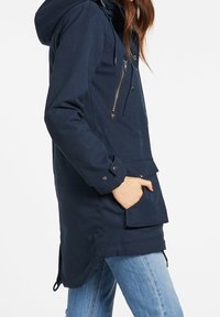 Mujer con chaqueta con capucha de color azul marino con cremallera lateral y bolsillo frontal con solapa, con la mano dentro del bolsillo, combinada con jeans de color azul claro.