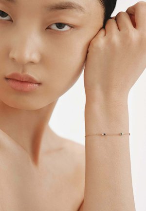 Pulsera de oro con dos pequeñas piedras preciosas de colores—una azul y una verde—unidas a una delicada cadena, llevada en una muñeca. Textura de piel suave.