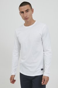 Langemouwig, effen wit shirt van katoen. Heeft een ronde hals en een subtiel merklabel aan de onderzijde. Soepele textuur.