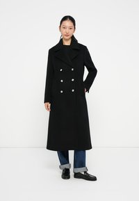 Manteau noir en laine croisé avec de grands boutons en argent, col cranté, poches latérales et une silhouette longue et droite. Porté sur un jeans bleu.
