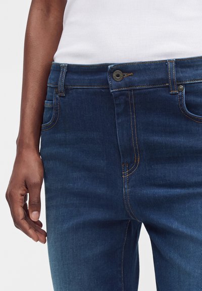 Jean en denim bleu foncé avec surpiqûres visibles porté par une personne portant un débardeur blanc côtelé, montrant la taille et la partie supérieure de la cuisse.