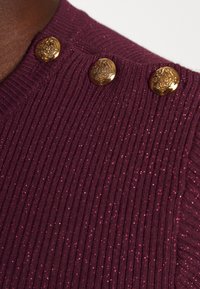 Bordeauxrode geribbelde trui met metalen gouden knopen, met een subtiele glinsterende textuur en golvende randen bij de halslijn.