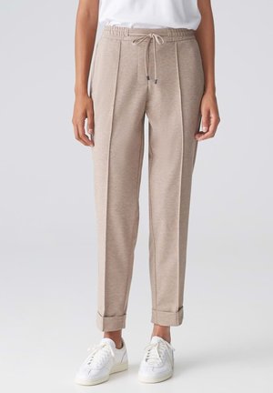 Beige snørrebukser med frontsømme og op rullede manchetter, båret med hvide sneakers og en hvid top mod en ensfarvet baggrund.