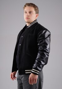 Schwarze Collegejacke mit Lederärmeln, gestreiften Rippbündchen und Knopfverschluss vorne. Sie verfügt über ein gesticktes Logo auf der Brust.
