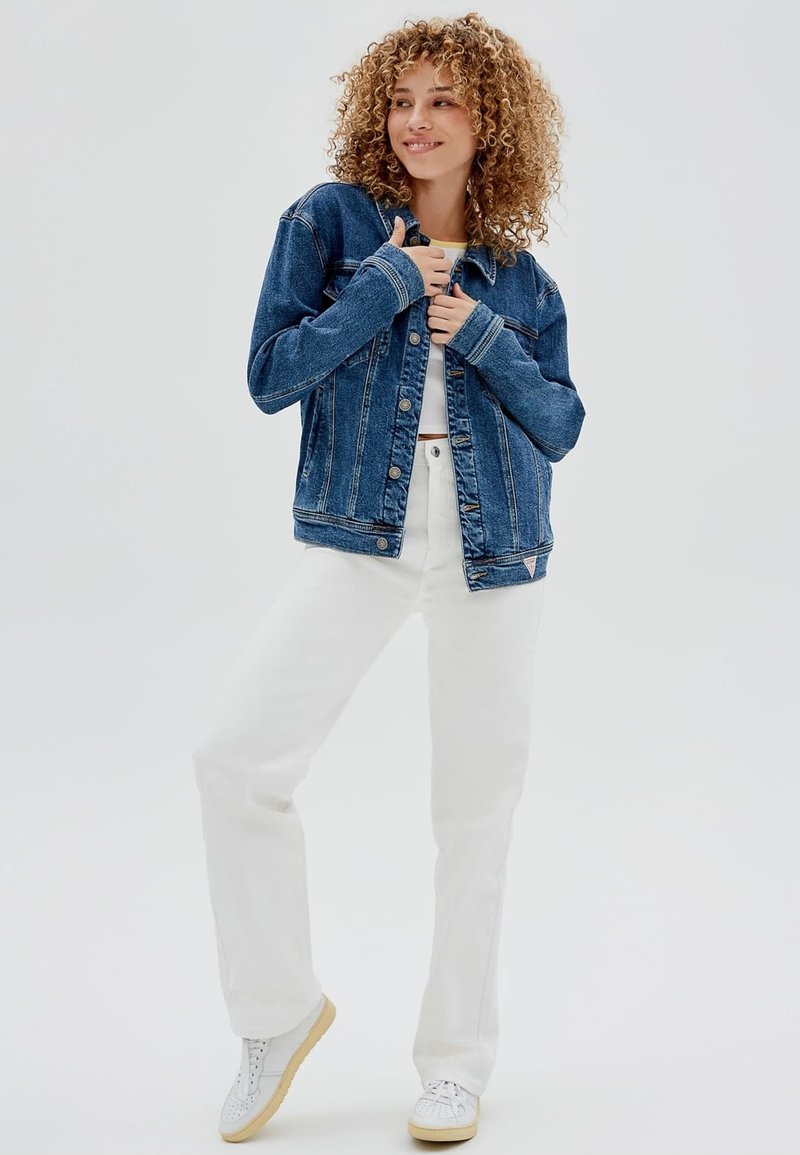 Giubbotto in denim blu scuro con chiusura a bottoni, dotato di tasche laterali e cuciture a contrasto, abbinato a pantaloni bianchi e sneakers bianche.