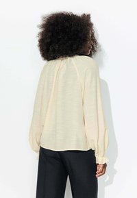 Blouse beige à manches longues avec détail de col froncé et poignets élastiques, associée à un pantalon noir, mettant en valeur un tissu doux et texturé.